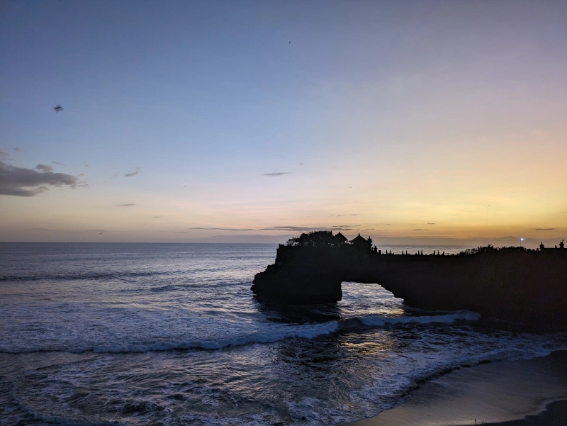 Atardecer Tanah Lot Formación rocosa en forma de arco que se adentra en el mar con el atardecer al fondo, vista desde el templo Tanah Lot de Bali
