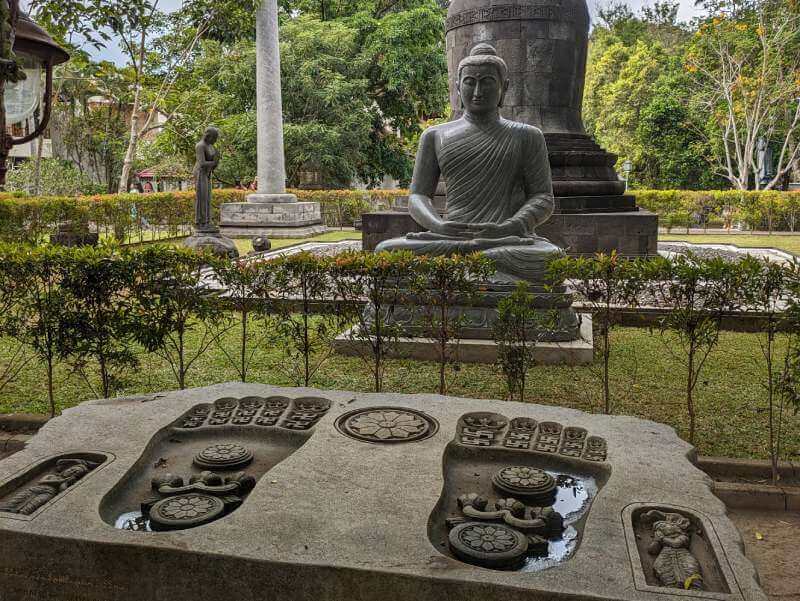 Piedra con con dos huellas de pies marcadas y varios simbolos representativos, enfrente de una estatua de buda en el monasterio Mendut