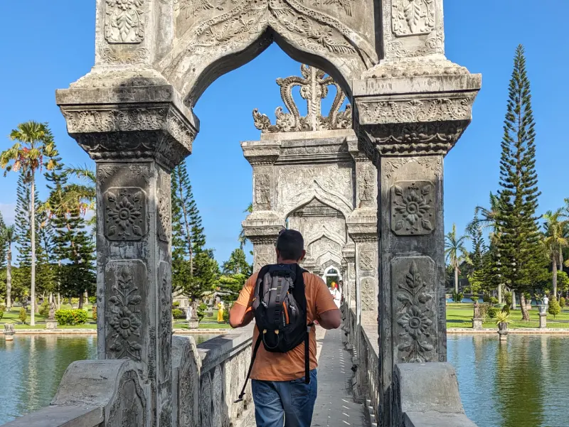 Jordi de espaldas cruzando un puente de piedra decorado con tallados florales en el palacio Taman Ujung.