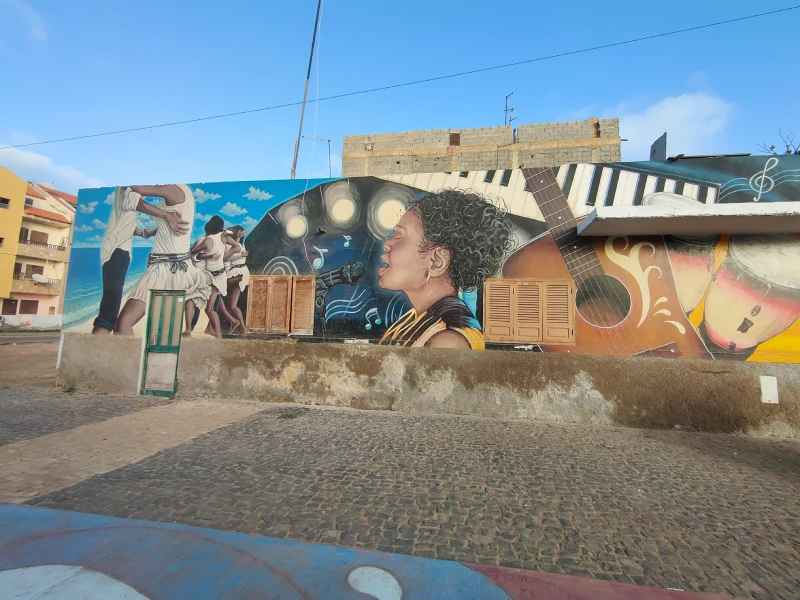 Mural musica Santa Maria Mural pintado en una pared, que representa una mujer negra cantando (posiblemente Cesaria Evora), una guitarra y varias personas bailando junto a la playa