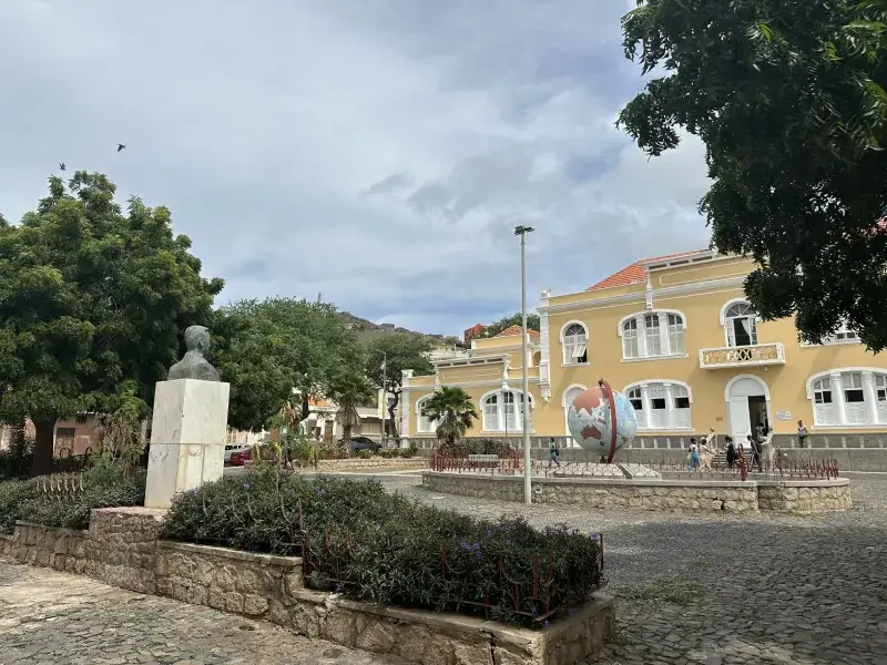 Universidad de Mindelo Edificio amarillo y blanco situado enfrente de una plaza con una bola del mundo en el centro y una escultura justo delante, rodeada de árboles